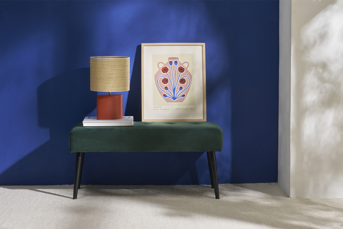 Banc GUESTA en velours vert avec pieds en m�tal, d�cor� d'une lampe et d'un cadre, devant un mur bleu.