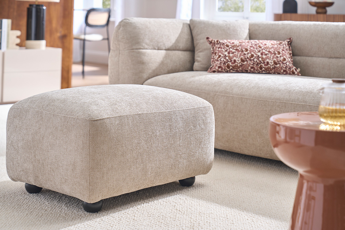 Pouf repose pieds en tissu beige, avec canap� en arri�re-plan, vue de 3/4